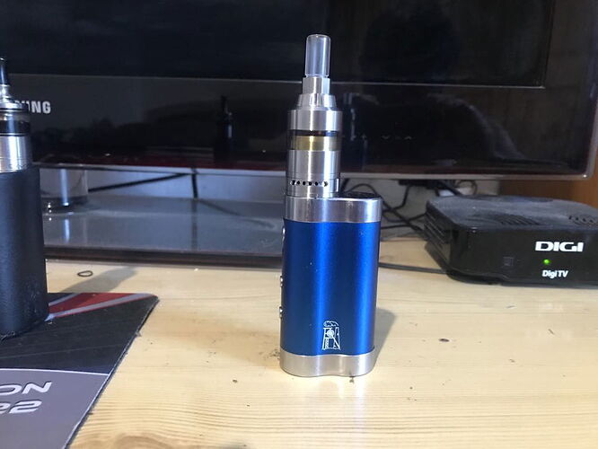 Az asztalon egy kék színű elektromos cigaretta (vape) áll, mögötte egy televízió és egy DigiTV set-top box látható. (Feliratozva AI-val)