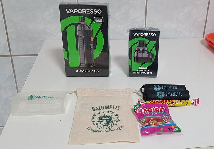On the table are two Vaporesso e-cigarette boxes, a cotton pouch with Calumette logo, two empty batteries, a plastic holder, and some candies. (Captioned by AI)