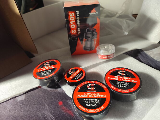 Divers accessoires de vapotage tels que des bobines de fil et une boîte Solo 2 RTA disposés sur une surface à motifs. (Légendé par IA)