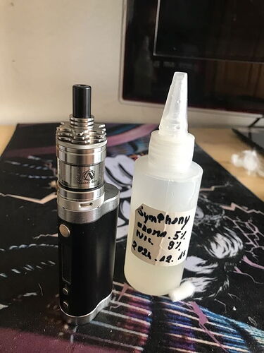 Az asztalon egy fekete-ezüst elektromos cigaretta és mellette egy kézzel írt címkével ellátott e-liquid flakon látható. (Feliratozva AI-val)