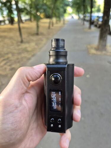 A hand holding a black electronic cigarette (vape) in a park, where trees and a road are visible in the background. (Labeled by AI)
