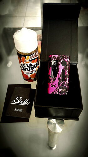 A képen egy fekete dobozban egy rózsaszín és fekete mintás elektronikai eszköz, mellette egy Slubby márkájú használati útmutató és egy De La Crump dohányízű folyadék található. (Feliratozva AI-val)