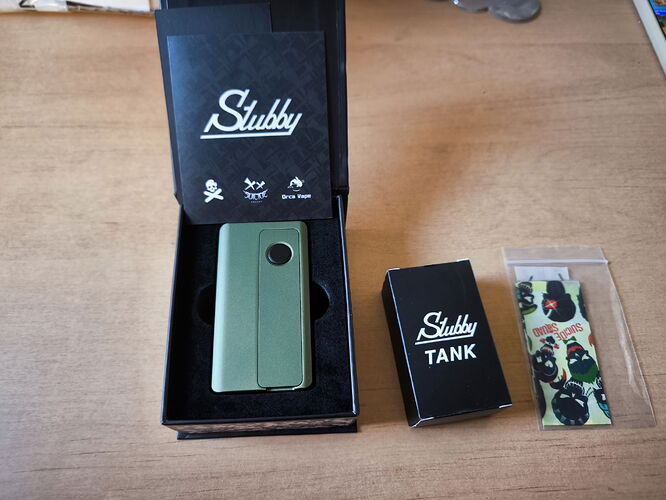 A green device is shown in the image, inside a black box, next to a small box labeled "Stubby Tank" and a pouch that likely contains stickers. (Autogenerated by AI)