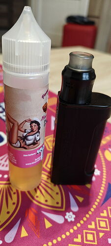 A bottle of e-liquid decorated with a female pin-up pattern and a black e-cigarette stand on a colorful tablecloth. (Captioned by AI)