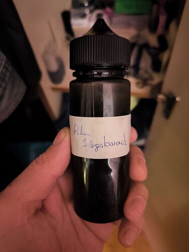 Une bouteille en plastique noir tenue dans une main, avec une étiquette écrite à la main indiquant "Cuisine de pruneaux". (Légendé par l'IA)