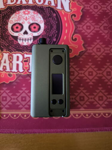 The image shows a gray device with a display and buttons on the side, on an ornate mat with a Mexican-style pattern. (Autogenerated by AI)