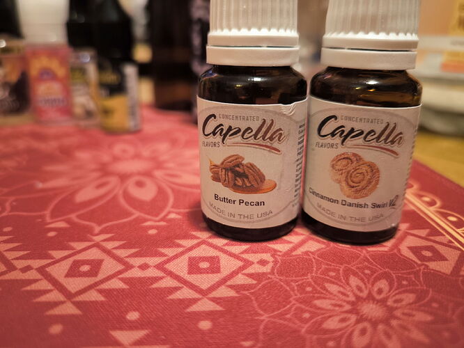 Two Capella aroma bottles are visible on a red, patterned tablecloth, one Butter Pecan and the other Cinnamon Danish Swirl flavor. (Labeled by AI)