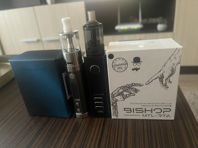 Two electronic cigarettes and a white box are visible on the table, featuring drawn hand heads and the text "BISHOP MTL-RTA". (Labeled by AI)