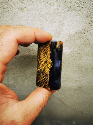 Une main tenant un objet petit, cylindrique, en bois et en matériau sombre devant un fond gris. (Légendé par IA)