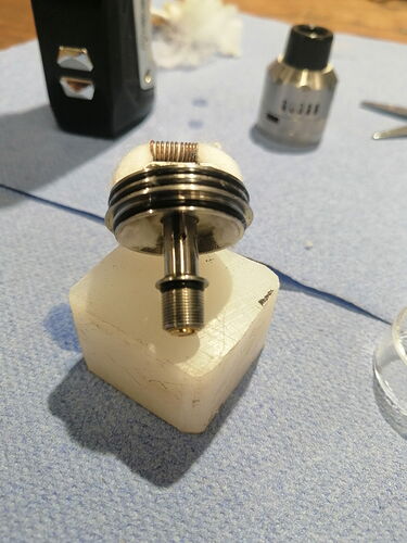The image shows a metal part, likely part of a vape device, placed on a white sponge block, with various tools in the background. (Caption by AI)