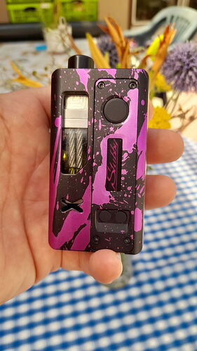 A hand is shown holding a pink and black splatter-patterned vape device, with blurred flowers and a checkered tablecloth in the background. (Labeled by AI)