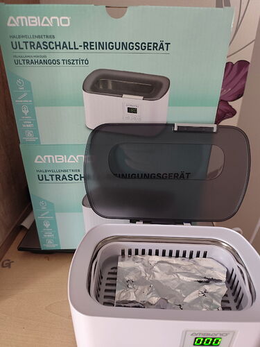 Az ultraszónikus tisztítógép nyitott fedéllel, benne alumínium fóliával és a doboza hátterében látható. (Feliratozva AI-val)