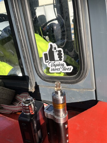 Az ablakon egy Vaping saves lives feliratú matrica látható, míg előtte két elektromos cigaretta áll egy piros felületen. (Feliratozva AI-val)