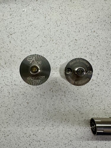 Az képen két különböző méretű és formájú vape alkatrész látható egy sima, világos felületen. (Feliratozva AI-val)