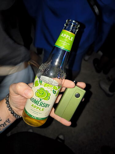 Une main tenant une bouteille de cidre Somersby saveur Pomme, tout en tenant un smartphone. (Légendé par IA)