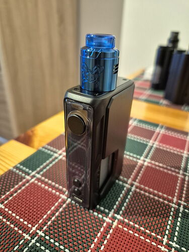 In the picture, a black multi-functional vape device is visible, with a blue cap on top, on a checkered tablecloth. (Labeled by AI)