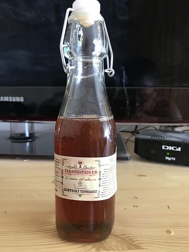 Une bouteille de liquide brun foncé est posée sur une table en bois, avec une étiquette dessus TABACCHIPICIO 3.0, un téléviseur et un récepteur Digi TV sont visibles en arrière-plan. (Légendé par IA)