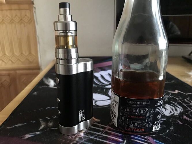 Az képen egy fekete és ezüst elektromos cigaretta látható egy üres palack mellett, amely valószínűleg dohányféléket tartalmazott. (Feliratozva AI-val)