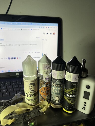Sur la table se trouvent quatre flacons d'e-liquide, un appareil e-cigarette blanc et une dragonne colorée devant un ordinateur portable. (Légendé par IA)