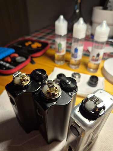 Két fekete és egy fehér vape mod áll az asztalon, melyek felső részén tekercsek láthatók, háttérben pedig folyadékpalackok és egy szerszámos doboz található. (Feliratozva AI-val)