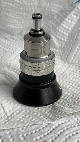 The image shows a metal and black plastic component, likely an industrial or technological device, standing in front of a white, textured paper background. (Captioned by AI)