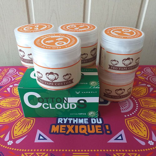 Ce qui est montré sur cette image, ce sont plusieurs boîtes de coton Pro Vape Cotton, placées sur une boîte Cotton Clouds vert-rouge, ainsi que sur un sous-verre coloré au motif de style mexicain. (Légendé par IA)