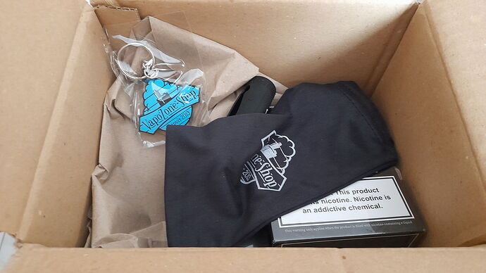 An open cardboard box is visible containing a blue 'Vapozone Shop' keychain, a black bag with a 'Vape-Shop' logo, and a box with a nicotine warning. (Labeled by AI)