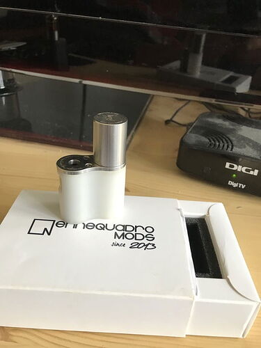 Az asztalon egy fehér "entequadro MODS" dobozon egy kis fém elektronikus eszköz látható, háttérben egy televízió és egy Digi TV vevő. (Feliratozva AI-val)