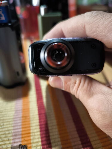 Egy kéz egy kis kamerát vagy videofelvevőt tart, melynek lencséje közelről látható. (Feliratozva AI-val)