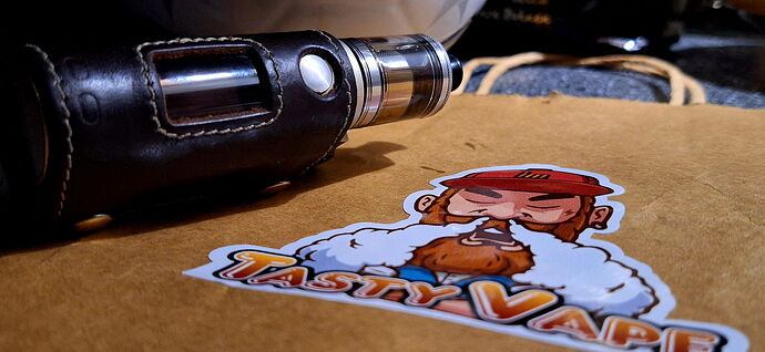 Une cigarette électronique repose dans un étui en cuir noir sur un sac en papier brun, avec une figure barbu et l'inscription "Tasty Vape". (Légendé par IA)