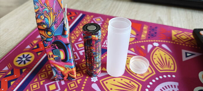 Boîtier coloré et à motifs, contenant une cigarette électronique et un support en plastique transparent sur une nappe à motifs. (Sous-titré par IA)