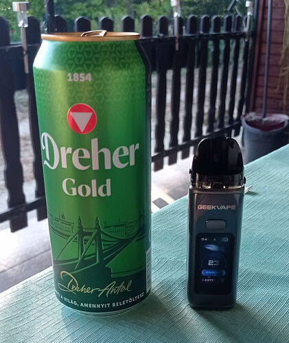 The picture shows a green Dreher Gold beer can and a black GeekVape electronic cigarette on a green table, with a fence and plants in the background. (Caption by AI)