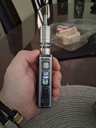 A hand holds a black vape mod with a screen, with a piece of cake in a plastic box visible in the background. (AI-generated caption)