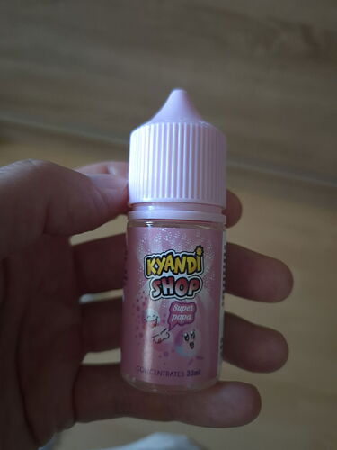 Une main tient une petite bouteille de liquide de la marque Kyandi Shop, avec une étiquette rose. (Légendé par l'IA)