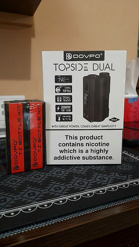 La photo montre la boîte d'un Dovpo Topside Dual Vaporizer et deux flacons rouges Dovpo 45 ml Flüssig Bottle Kit sur une table noire. (Légendé par IA)