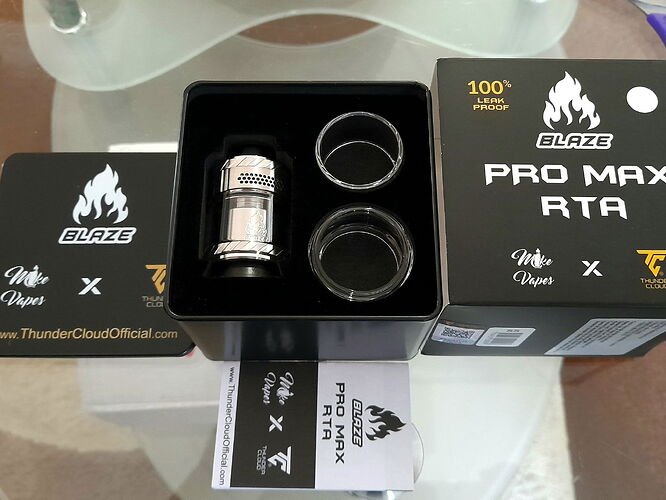 Az asztalon egy BLAZE PRO MAX RTA vape tartozék készlet van kinyitva, benne egy fém atomizátor és két üveg cső. (Feliratozva AI-val)