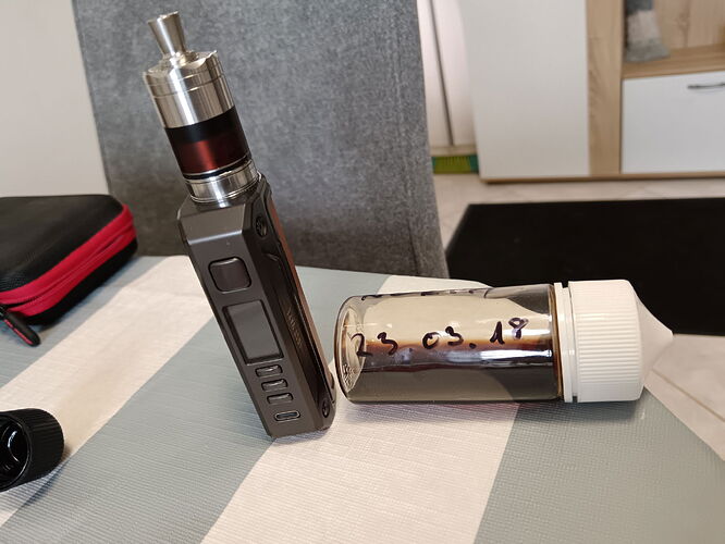 Un appareil de vape noir et un récipient blanc à bouchon sur une table, sur lequel on voit de petits ingrédients ou accessoires. (Légendé par IA)