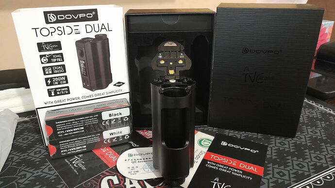 La photo montre un appareil de cigarette électronique DOVPO TOPSIDE DUAL, sa boîte et ses manuels d'utilisation. (Légendé par IA)