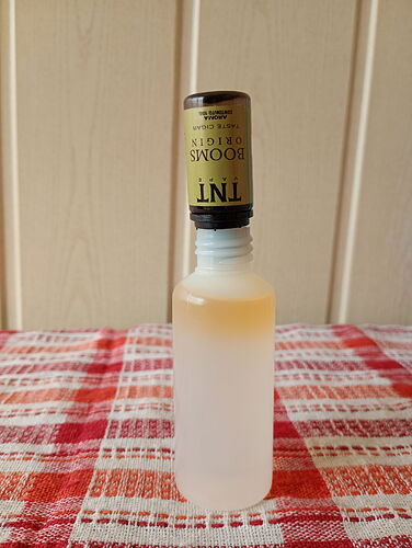 A képen egy átlátszó műanyag palack látható, amelynek nyaka fehér, és egy felfordított T.N.T. Brooks Originals doboz van a tetején, a háttérben pedig egy világos fal és egy piros-fehér kockás terítő. (Feliratozva AI-val)