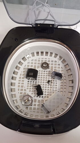 Egy ultrahangos tisztító gép nyitott fedelével és szétbontott apró fémdarabokkal a belsejében. (Feliratozva AI-val)