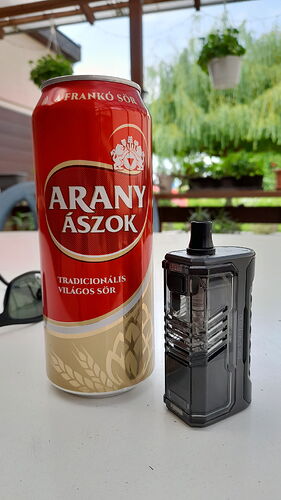 Une canette de bière Arany Ászok et une cigarette électronique noire sont posées sur une table blanche, avec des plantes et une cour visibles en arrière-plan. (Légendé par IA)