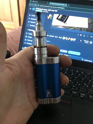 Egy kéz egy kék és ezüst színű elektromos cigarettát tart egy laptop előtt, amelyen egy internetes fórum vagy közösségi oldal látható. (Feliratozva AI-val)