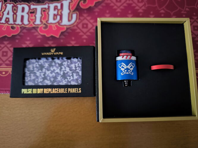 Az képen egy Vandy Vape Pulse BF RDA vape tartozék és egy dobozban lévő kék, koponyás vape alkatrész látható cserélhető panelek mellett. (Feliratozva AI-val)