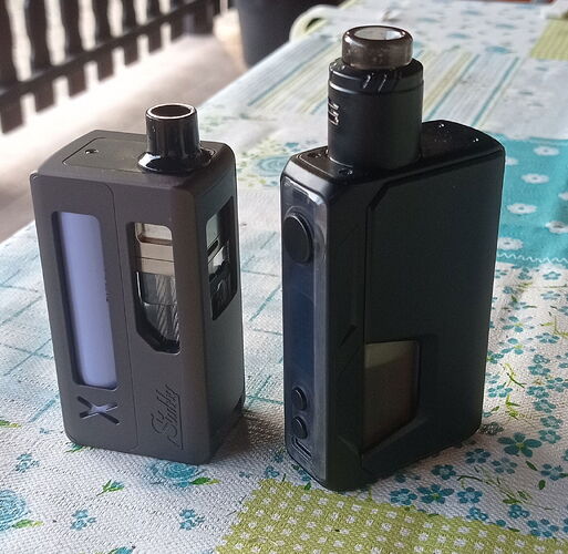 Két különböző fekete elektromos cigaretta eszköz áll egy virágmintás asztalterítőn. (Feliratozva AI-val)