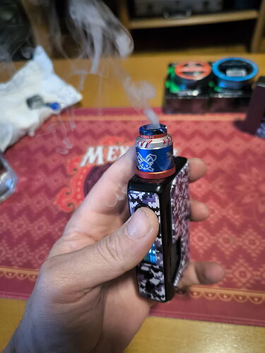 Egy kézben tartott elektromos vaporizerből gőz száll fel egy piros szőnyegen lévő asztal fölött. (Feliratozva AI-val)