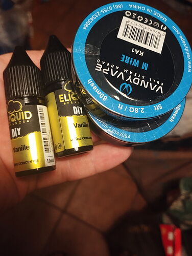 Deux petits flacons DIY avec des étiquettes jaunes et deux bobines Vandy Vape M Wire avec des bouchons bleu et noir sont tenus dans une main. (Légendé par IA)