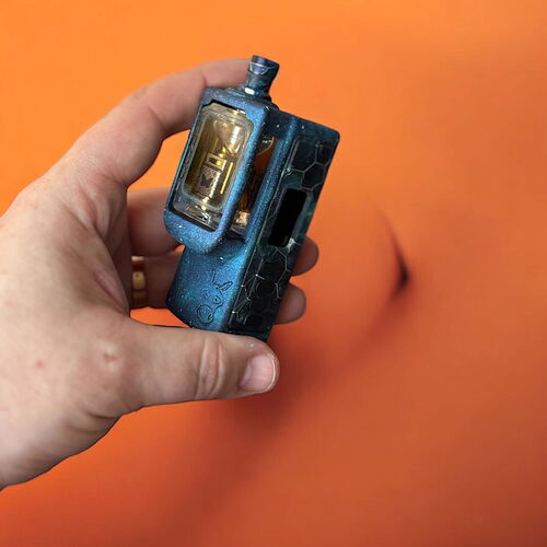 A képen egy kézben tartott, kék ízű vape látható, amelynek a közepén egy átlátszó tartály található, míg a háttérben egy narancssárga felület van. (Feliratozva AI-val)