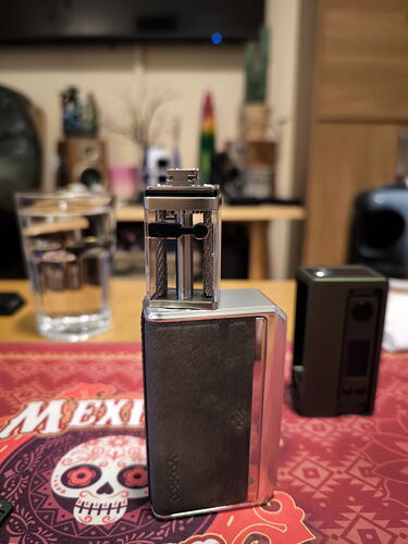 Ezen a képen egy ezüstszínű vape eszköz látható, amely egy piros mintás terítőn áll, háttérben egy víglassal. (Feliratozva AI-val)