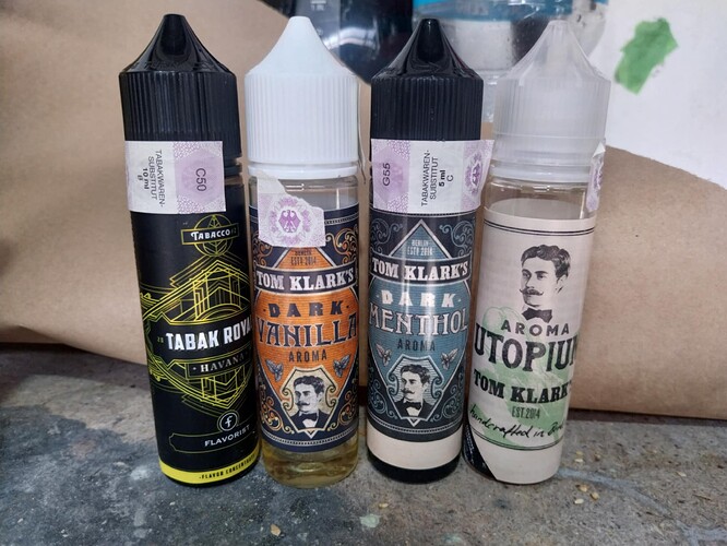 Quatre bouteilles d'e-liquide sont visibles sur la photo, parmi lesquelles figurent les arômes TABAK ROY, DARK VANILLA, DARK MENTHOL et UTOPIUM de la marque Tom Klark's. (Légendé par IA)