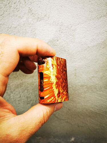 Une main tenant un petit objet carré marron à motif devant un mur clair et lisse. (Légendé par IA)